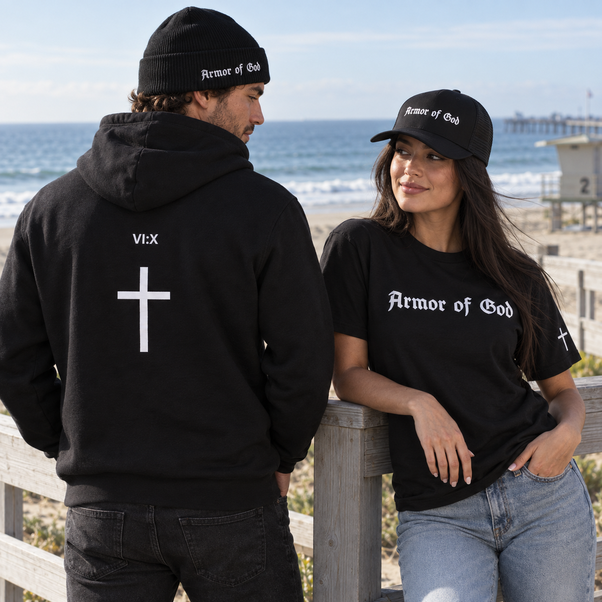 Armor of God cuffed beanie and cross-back hoodie beside a woman wearing the trucker cap and Classic T-Shirt at the coast